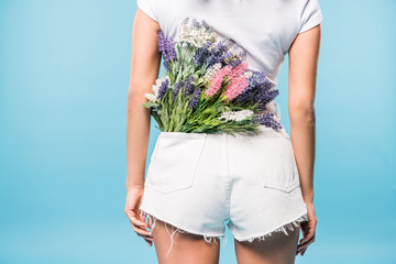 Cropped view of woman in shorts with flower bouquet in pocket on blue background