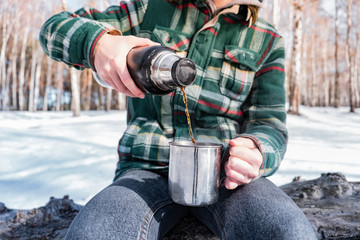 Pouring hot drink out of thermos at a campsite. Person in a winter forest during a hiking trip getting warm