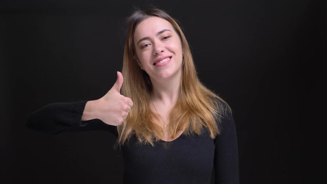Closeup portrait of young beautiful caucasian female showing thumb up and smiling while looking at camera with background isolated on black