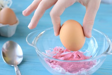 White woman fingers holding brown easter egg above the glass bowl with red foam dye. Traditional Easter dyeing eggs on blue wooden table with blurred egg and spoon. Homemade Easter eggs painting
