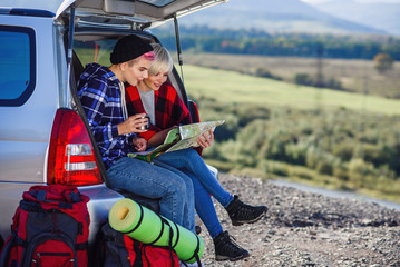 Young friends travelers sitting on trunk of car with cap of tea and looking at the paper map. Two happy hipster girls enjoying on vacation in mountains. Summer travel concept.