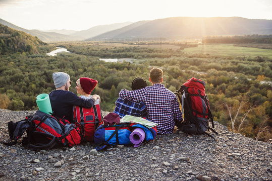 Group of young friends traveling together in mountains. Happy hipster travelers with backpacks sitting on the top of mountain at sunset background. Traveling, tourism and friendship concept.