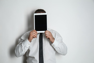 Businessman hiding face behind tablet computer on light background