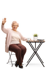 Senior woman sitting at a table with a cup of coffee and greeting with hand