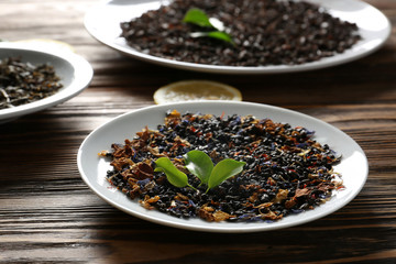 Plates with different types of dry tea leaves and lemon slices on wooden table