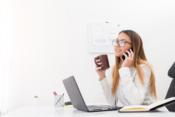 Cheerful businesswoman with eyeglasses holding a coffee cup while talking on mobile in her office.