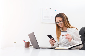 Attractive young businesswoman buying with a bank card on mobile phone while sitting in her office.
