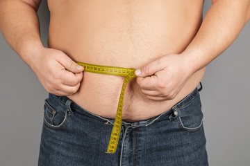 A man measures his fat belly with a measuring tape. on a gray background.