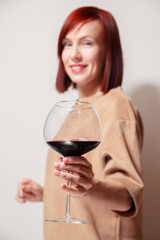 Young beautiful redhaired woman with bright lipstick on white background is smiling and holding a glass of red wine at professional blind tasting sommelier competition