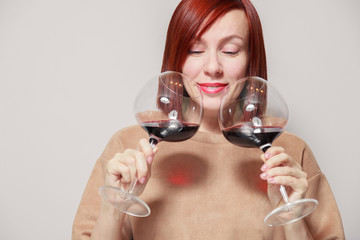 Young cheerful red haired girl with bright lipstick on a white background holds and sniffs two glasses of red wine and assesses aroma and notes at a professional blind tasting contest