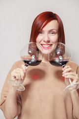 Young cute smiling red haired girl with bright lipstick on a white background holds and sniffs two glasses of red wine and assesses the aroma and notes at a professional blind tasting contest