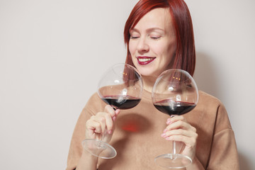 Young cute smiling red haired girl with bright lipstick on a white background holds and sniffs two glasses of red wine and assesses the aroma and notes at a professional blind tasting contest