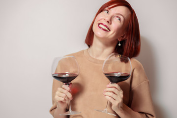 Young cheerful redhaired girl sommelier on white background smile and hold with two glasses of red wine. Concept alcoholism, alcohol addiction, bad habit
