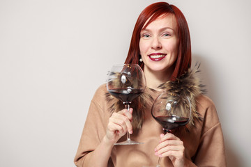 Young funny redhaired girl sommelier with fur cape on white background smile, hold two glasses red wine. Concept alcoholism, addiction, bad habit, collection great rare wines, winelover, expert