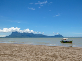 A beach in Bako National Park in Borneo, Malaysia. Yellow sand mixing with algae on the ground. Boat parked on the shore of the beach. High mountains in the back, covered with white clouds.