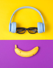 Minimalism flat lay concept. Smiling face listens to music. Headphones, 3D glasses, banana on purple yellow background. Top view