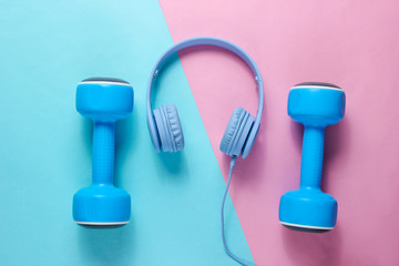 Blue headphones, plastic dumbbells on pink blue background. Sports with music. Flat lay. Top view.