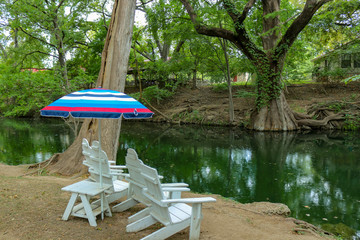 White adirondack lounge chairs and a colorful sun umbrella for relaxing on a summer day at Cypress Creek  a public park in Wimberley Texas