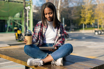 Young beautiful african american woman sitting outdoors in the city with a tablet pc while holding a take away coffee in a sunny day