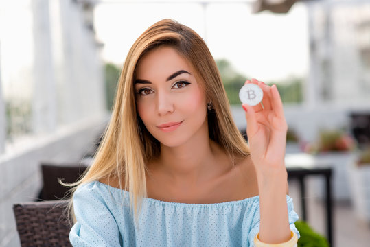 Beautiful girl in summer cafe. Tanned skin casual makeup, long hair, close-up of a woman in a blue dress. He holds a bitcoin cryptocurrency silver coin in his hand. A gentle smile on his face.