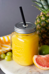 Pineapple cocktail or juice in glass jar bottle with straws and fruits on background - pineapple, oranges, grapefruit, lime and grapes. Gray background. Summer cold drink.