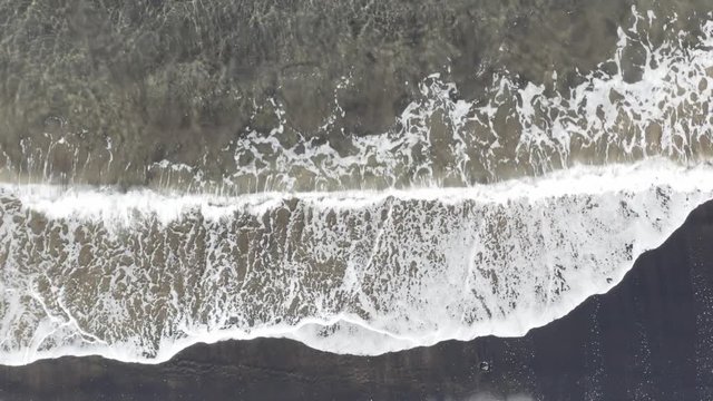 Drone shot pull back of peaceful waves roll into a black sand beach shore paradise