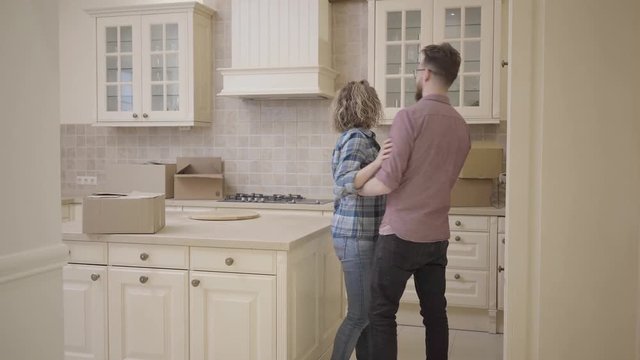 Bearded man and pretty woman enter the kitchen, holding hands. Man puts his wife on the table. Woman hugs her husband, they kissing. Married couple moves into a new home