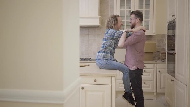 Bearded man stands near pretty woman sitting on table in big half empty kitchen. Woman hugs her husband, they kissing ans smiling. Married couple moves into a new home. Camera moves left behind wall