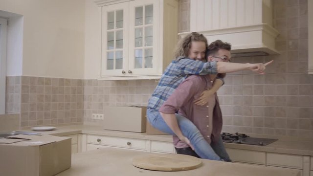Bearded man carries pretty woman sitting on his back in the kitchen. Woman hugs husband and shows him direction with her finger. Married couple have a fun at new home, unpacked boxes everywhere