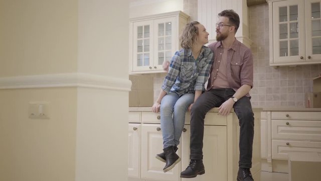Young kissing couple sitting on the kitchen table in a new apartment. Family moves to a new home. Bearded husband with glasses and a curly-headed wife with blond hair dressed in casual clothes.
