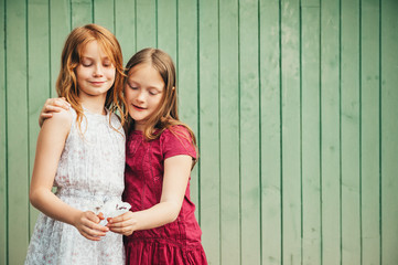 Outdoor portrait of two adorable little girls, wearing dresses, standing against green wooden...