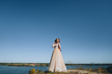 Fototapeta na wymiar A lovely bride in a beige dress with a crown is standing on a cliff, against a background of rocks and a blue sky. Wedding photography. Princess Bride.