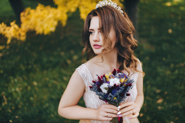 Fototapeta premium A smiling bride in a beige dress and a bouquet is standing in a park with yellow leaves. The princess with a crown enjoys the fall. Portrait of a beautiful bride with brown hair. Wedding photography.