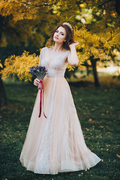 A beautiful bride in a beige dress is standing in a park with yellow leaves. The princess with a crown enjoys the fall. Wedding portrait of a cute bride with brown hair. Wedding photography.