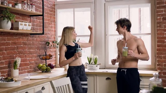 Attractive sport man with smoothie in hands showing his muscles on the arm to beautiful fit woman in the kitchen.