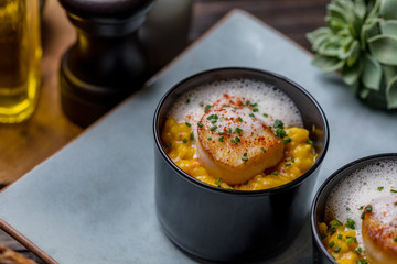Orange pumpkin risotto with scallop and foamy sauce in black bowl on rustic wooden table