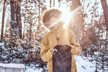 Young snowboarder dressed in warm clothes leans on a snowboard standing in a snowy forest during...