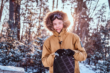 Young cheerful snowboarder dressed in warm clothes leans on a snowboard standing in a snowy forest...