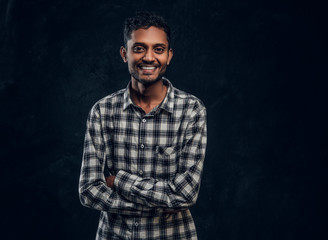 Portrait of a smiling Indian guy wearing a checkered shirt posing with his arms crossed and looking at the camera in a dark studio