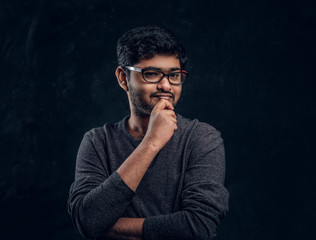 Young Indian guy wearing eyewear and casual clothes posing with a thoughtful look in a dark room