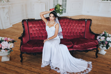 Obraz premium A beautiful bride in a white dress with a bouquet is sitting on a red sofa with flowers. Chic portrait of a cute and smiling brunette. Wedding photography. Floristry.