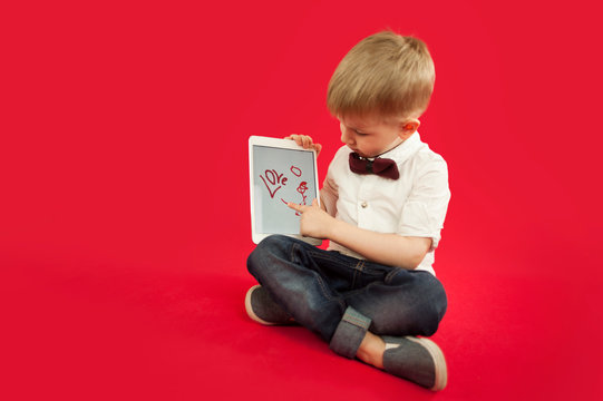 Child Boy Is Drawing In Tablet Ipad Red Heart On Red Background. Family And Love Relationship Concept