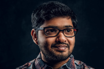 Close-up portrait of a young Indian guy in eyewear and checkered shirt in studio against the background of the dark wall
