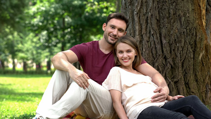 Happy pregnant family sitting under tree in park, courses for future parents