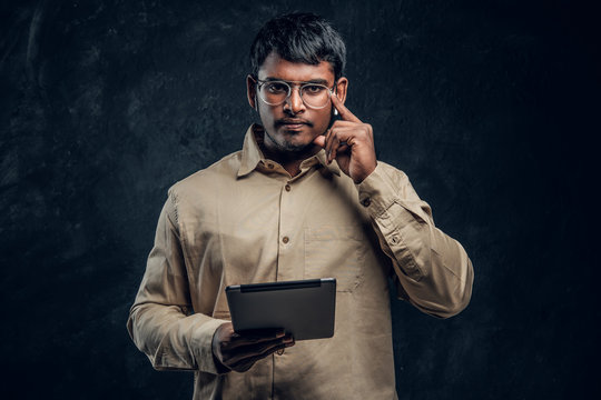Confident Indian man in eyewear and shirt holding a tablet computer and looking at a camera with a thoughtful look in dark studio