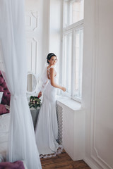 Fototapeta premium A beautiful bride in a white dress with a bouquet is standing near the window in a modern studio. Chic portrait of a cute brunette. Wedding photography.