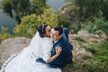 Newlyweds in love hugging against the background of rocks and a cliff. Portrait of a stylish groom with glasses and a cute bride in a white palette. Wedding photography.