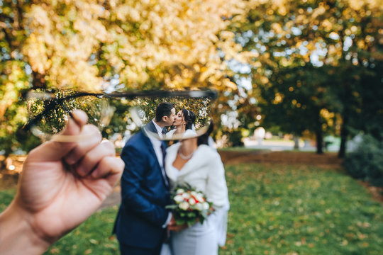 Lovers and beautiful newlyweds kiss through a lens in the autumn in the park with yellow leaves. Portrait of a stylish groom with glasses and a cute bride in a white dress. Wedding photography.