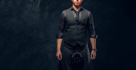 Handsome redhead man dressed in an elegant vest with tie and kilt in studio against a dark textured...