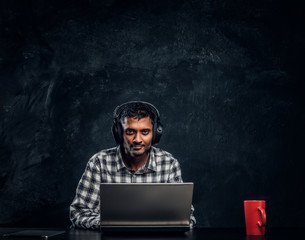 Indian student in headphones sitting at a desk with laptop and looking at a camera. Studio photo...